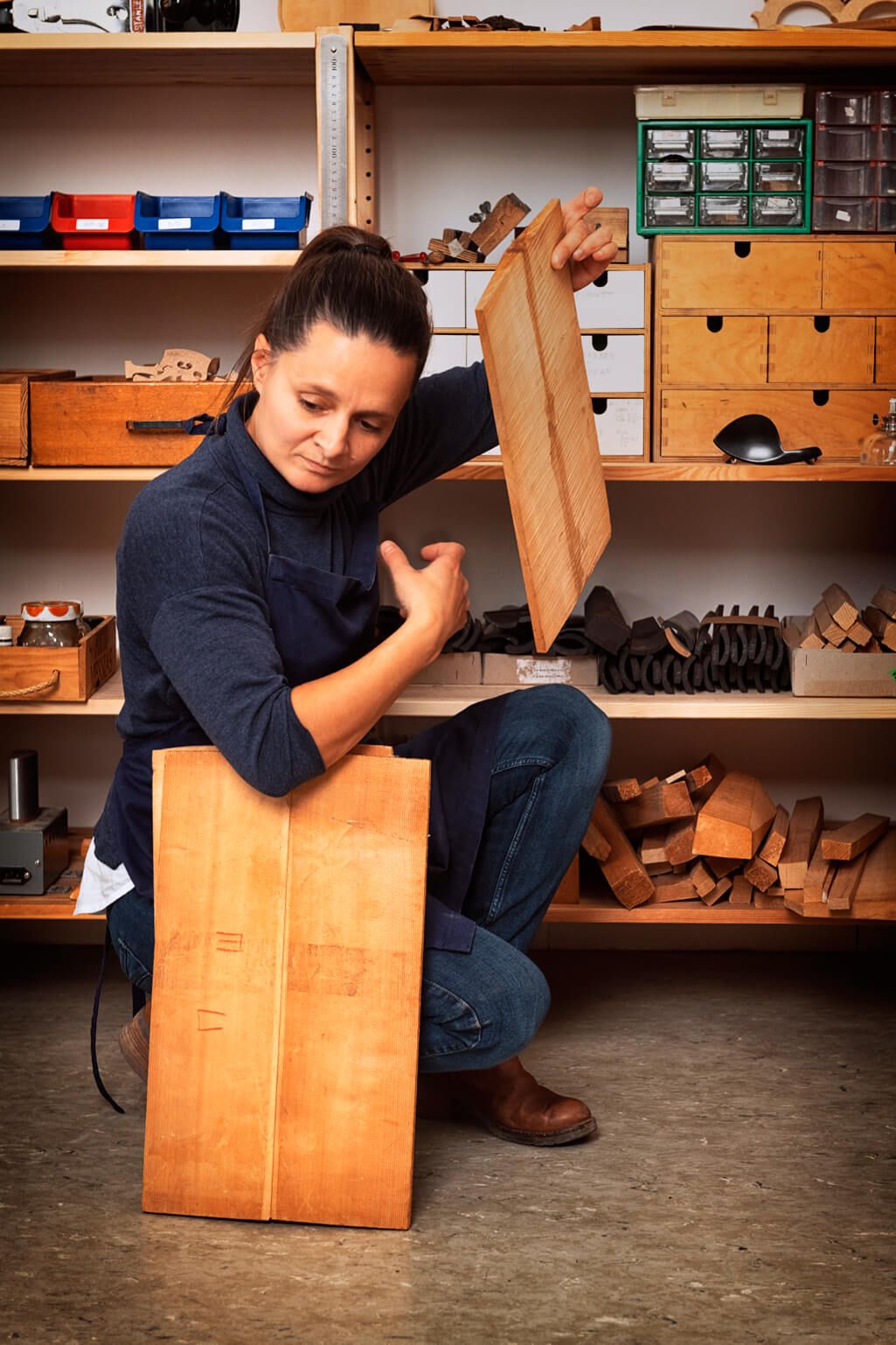 Judith Bauer kniet vor einem Regal mit verschiedenen Hölzern. Sie hält eine Platte mit der linken Hand vors Gesicht. Zwei weitere Platten hält sie unterm rechten Arm.