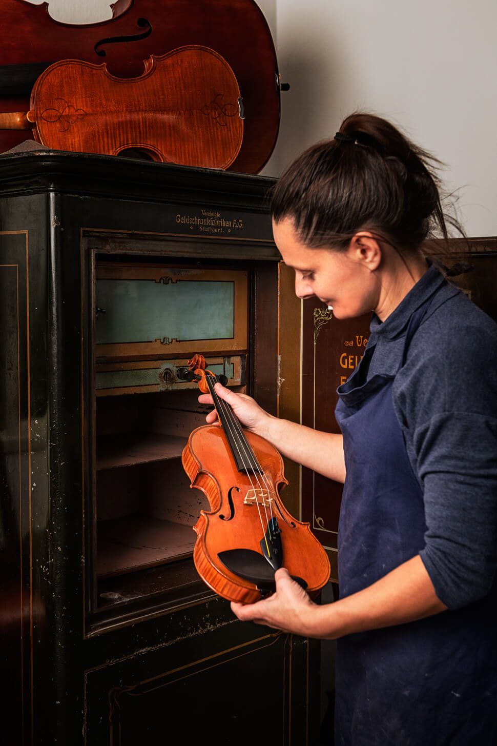 Judith Bauer steht vor einem altmodischen Geldschrank. Sie blickt auf eine Geige, die sie in der Hand hält.