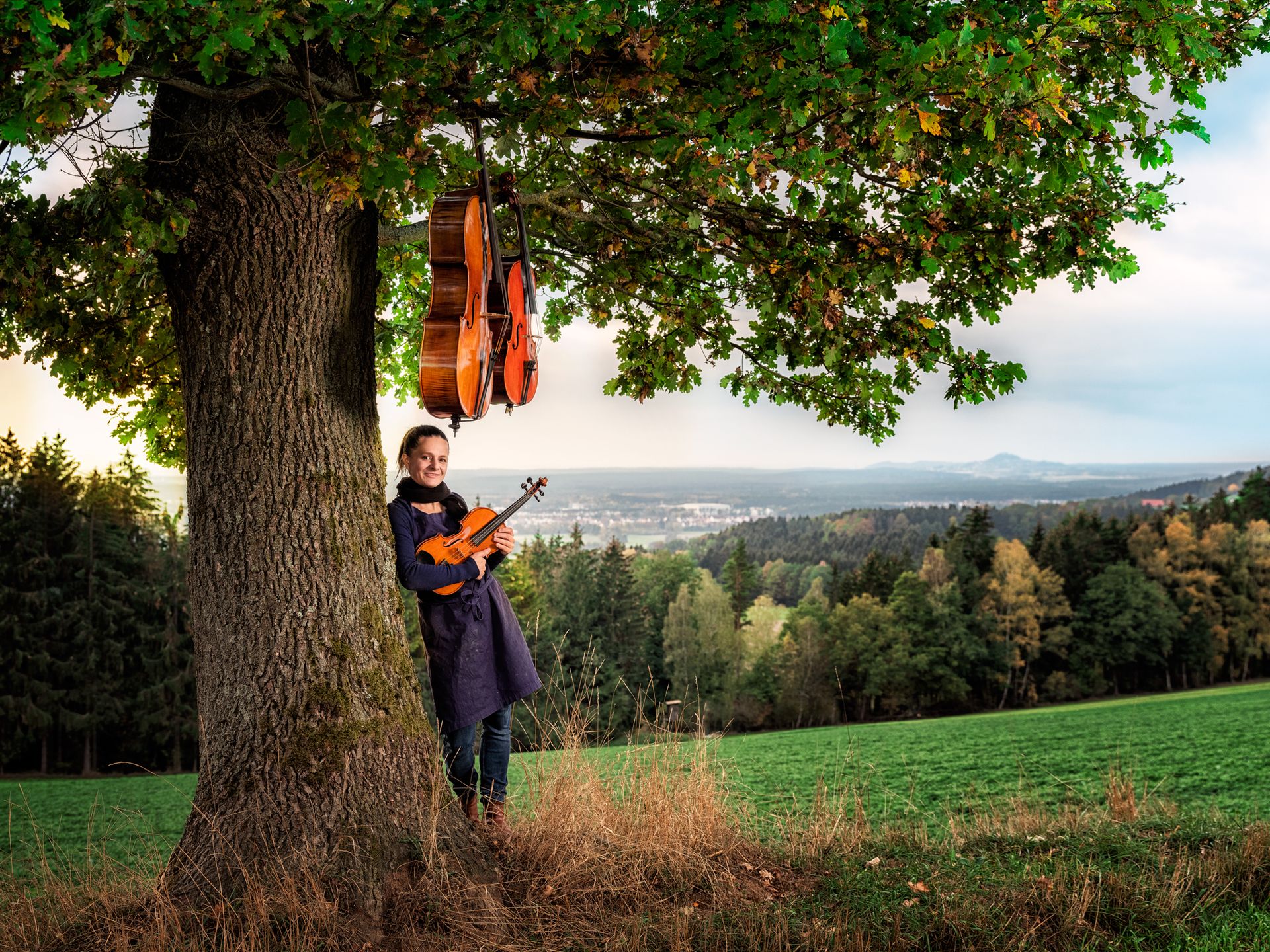Judith Bauer lehnt an einem Baum und hält eine Geige fest. In den Ästen des Baums hängen zwei Celli. Im Hintergrund liegen Wald und eine Stadt.