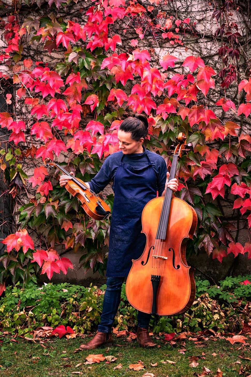 Judith Bauer steht vor einer Mauer, an der Weinlaub wächst. Sie blickt auf die Geige in ihrer rechten Hand. In der linken Hand hält sie ein Cello.