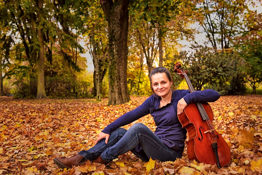Judith Bauer sitzt auf einer laubbedeckten Wiese. Im Arm hält sie ein Cello, im Hintergrund sind Bäume.